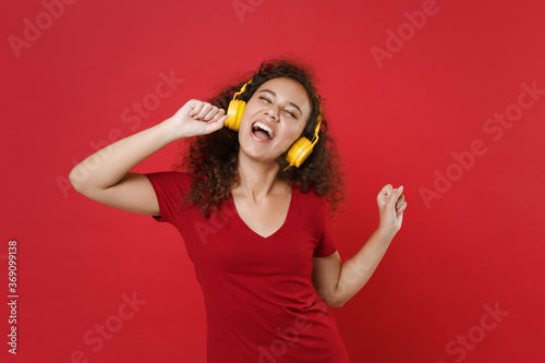 Funny young african american woman girl in casual t-shirt posing isolated on red background studio portrait. People lifestyle concept. Mock up copy space. Listening music with headphones, sing song.