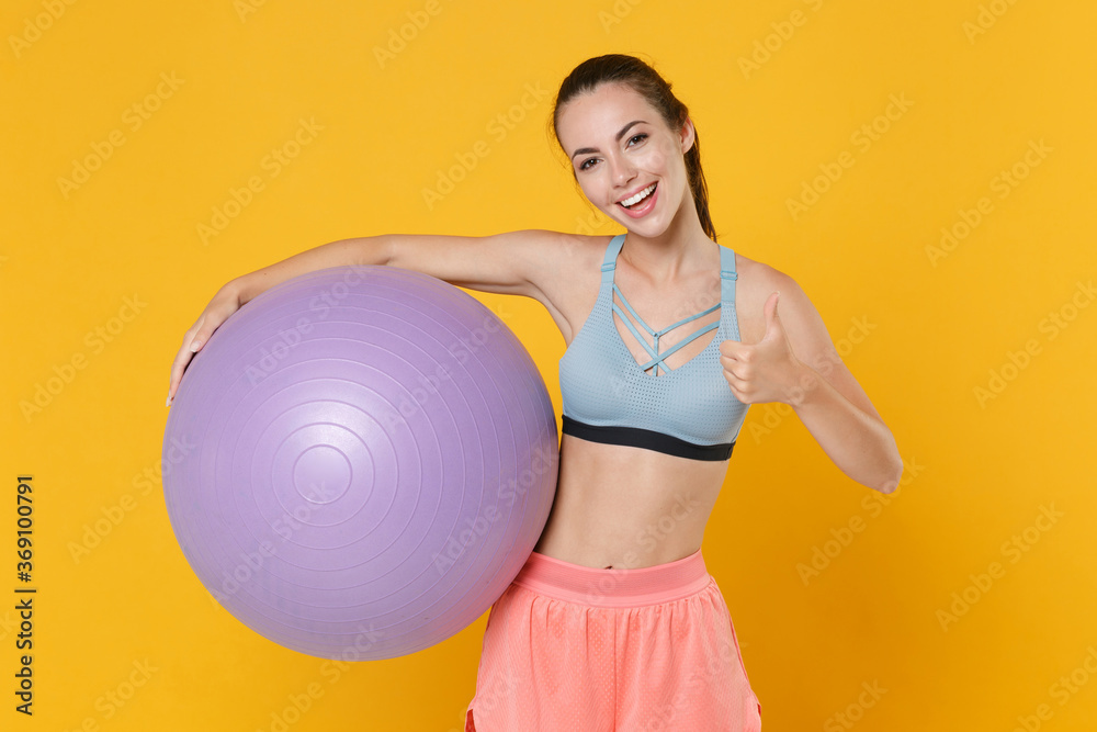 Cheerful young fitness sporty woman girl in sportswear working out isolated on yellow background studio portrait. Workout sport motivation concept. Mock up copy space. Hold fitball, showing thumb up. - obrazy, fototapety, plakaty 