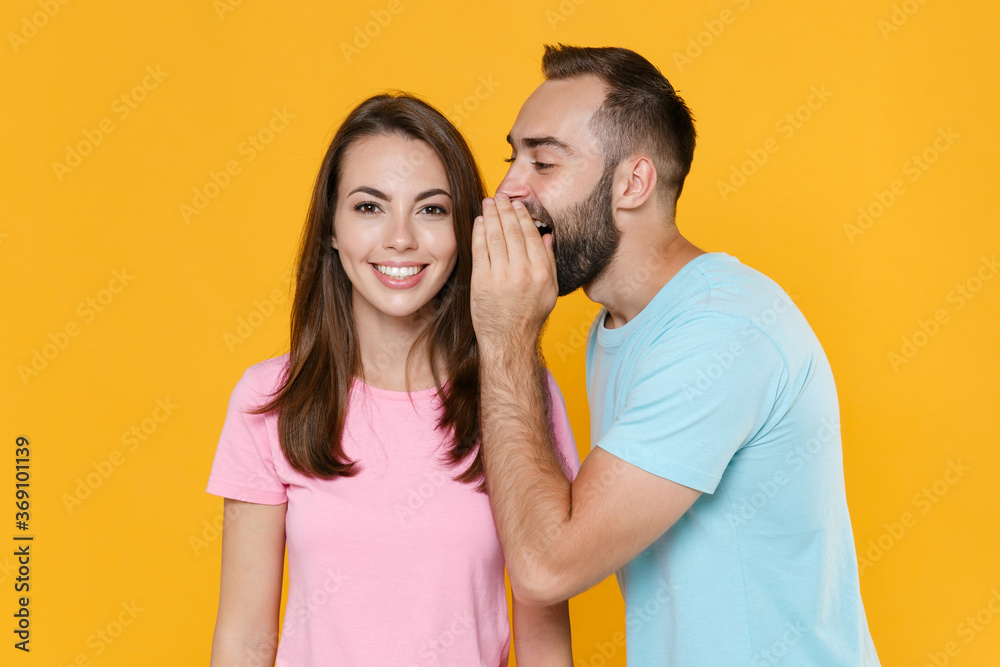Naklejka premium Smiling young couple friends guy girl in blue pink t-shirts isolated on yellow background studio portrait. People lifestyle concept. Mock up copy space. Whispering secret behind hand, sharing news.