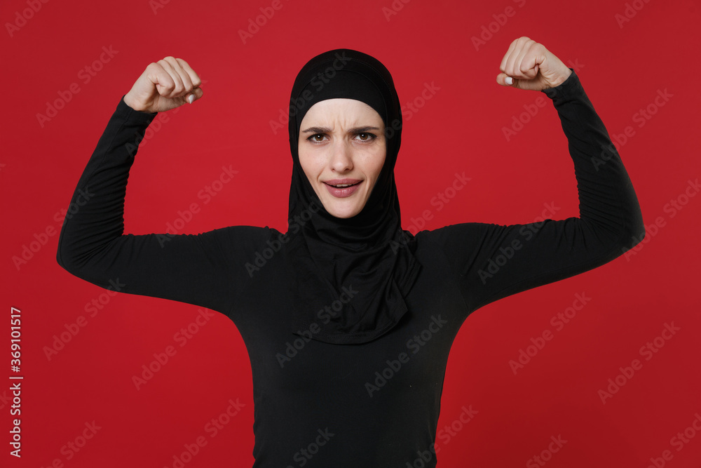 Strong young arabian muslim woman in hijab black clothes posing isolated on bright red wall background studio portrait. People religious lifestyle concept. Mock up copy space. Showing biceps, muscles. Strong young arabian muslim woman in hijab black clothes posing isolated on bright red wall background studio portrait. People religious lifestyle concept. Mock up copy space. Showing biceps, muscles.