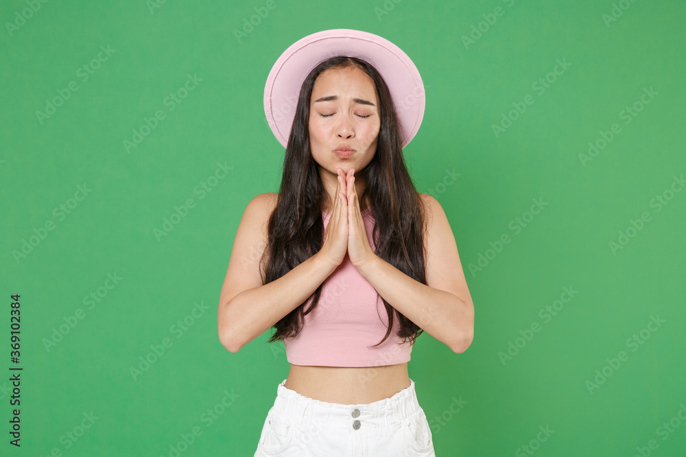 Pleading young asian woman girl in casual pink clothes hat posing isolated on green background studio portrait. People emotions lifestyle concept. Mock up copy space. Holding hands folded in prayer. Pleading young asian woman girl in casual pink clothes hat posing isolated on green background studio portrait. People emotions lifestyle concept. Mock up copy space. Holding hands folded in prayer.