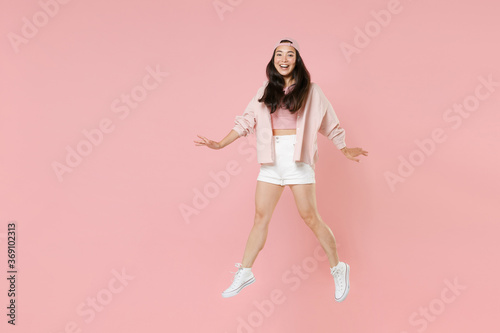 Full length portrait excited young asian girl in casual clothes, cap isolated on pastel pink background studio portrait. People lifestyle concept. Mock up copy space. Jumping spreading hands and legs.