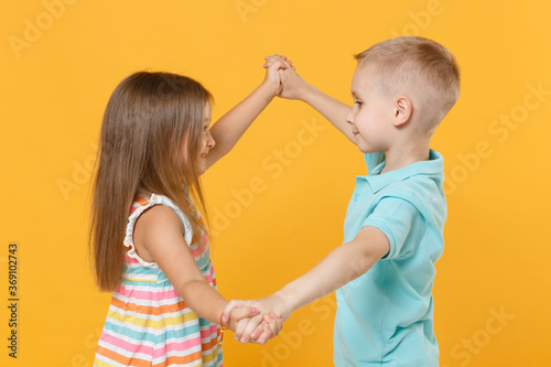 Little couple kids boy girl 5-6 years old in blue pink clothes shirt dress posing have fun isolated on yellow background children studio portrait. People childhood lifestyle concept hold hands dance