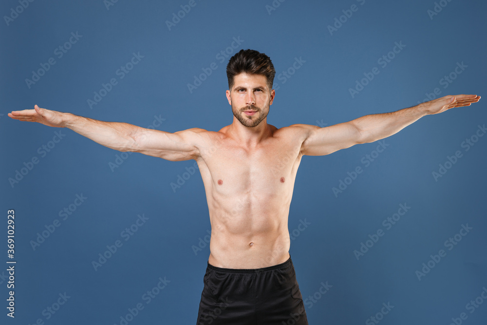 Handsome young bearded fitness sporty guy bare-chested muscular sportsman isolated on blue background studio. Workout sport motivation lifestyle concept. Posing with spreading outstretching hands.