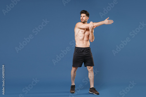Full length portrait of young bearded fitness sporty strong guy bare-chested muscular sportsman isolated on blue background. Workout sport motivation lifestyle concept. Doing stretching exercising. © ViDi Studio