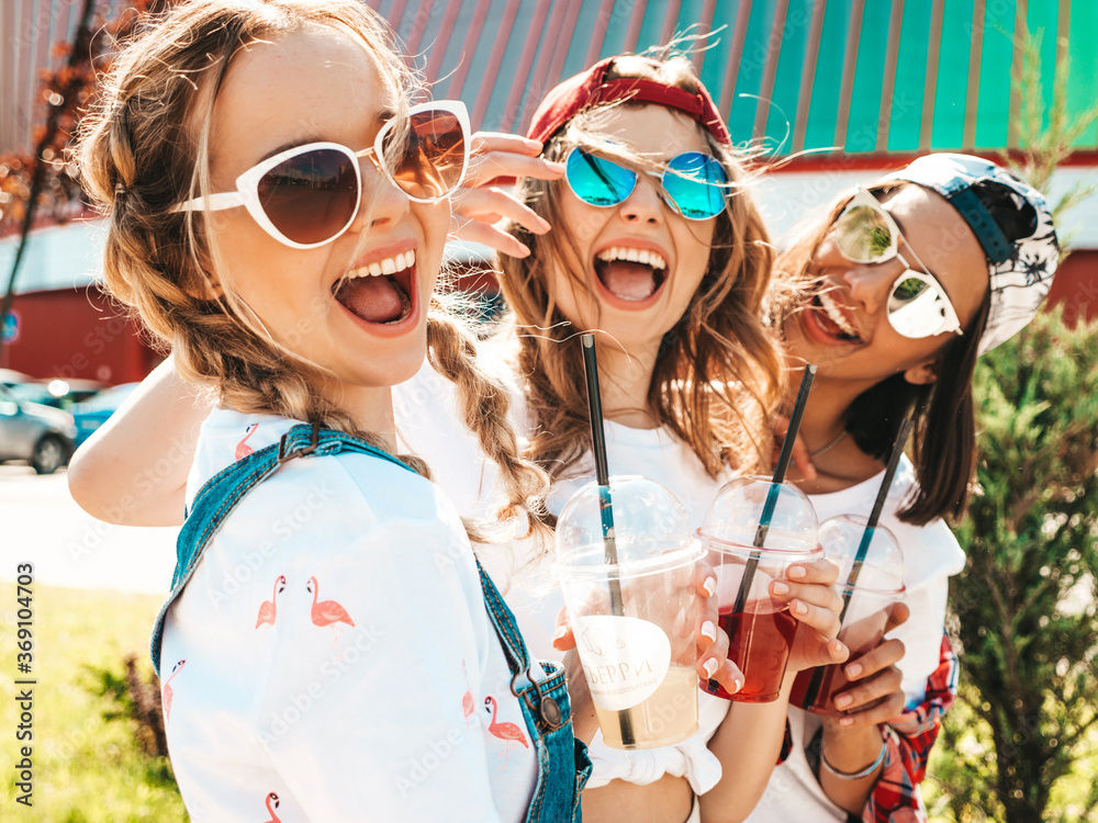 Three young beautiful smiling hipster girls in trendy summer clothes.Sexy carefree women posing outdoors.Positive models holding and drinking fresh cocktail smoothie drink in plastic cup with straw