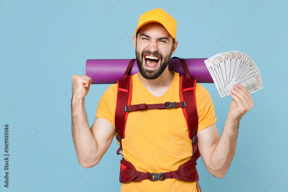 Happy traveler man with backpack isolated on blue background. Tourist traveling on weekend getaway. Tourism discovering hiking concept. Hold fan of cash money in dollar banknotes doing winner gesture. - obrazy, fototapety, plakaty 