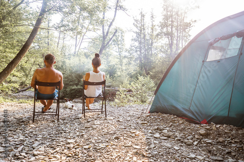 Relaxed couple sitting outside tent while camping nearby mountain river on a sunny day. Woman holding coffee talking with man. Two young caucasian people enjoying camping together.