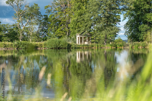 A view with blurred grass in the foreground across a beautiful lake with reflections of trees towards an old abandoned structure in the middle of a rural countryside scene