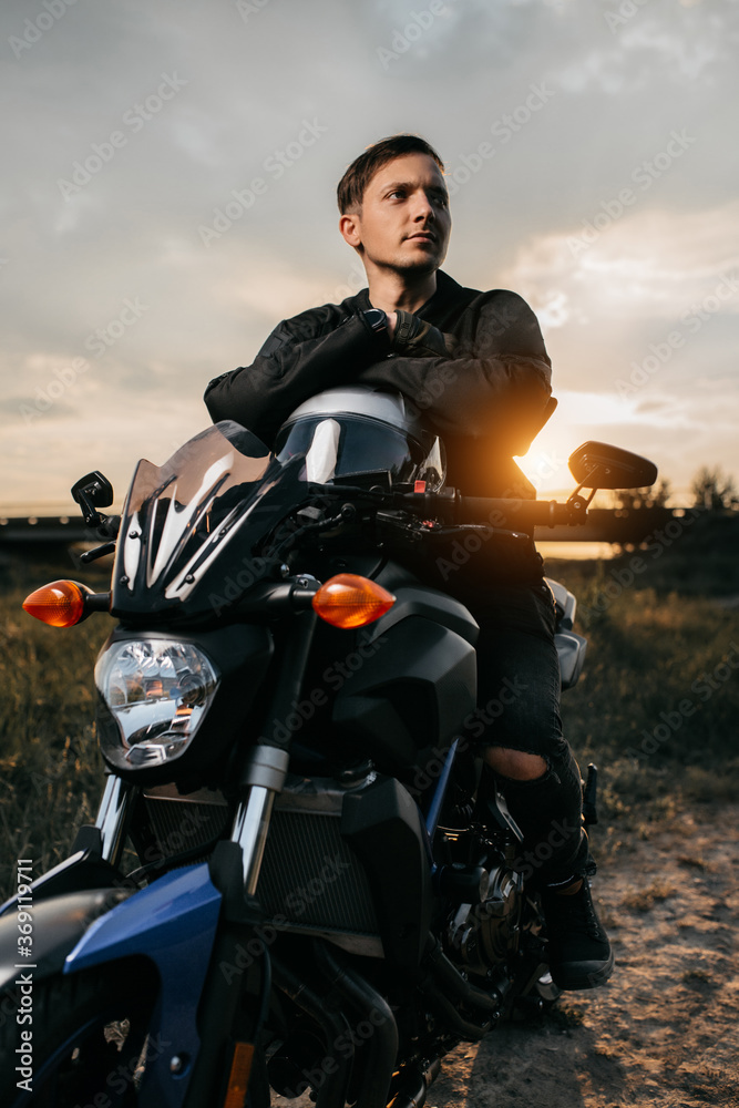 Close-up photo of handsome biker sitting on motorcycle in sunset on the country road. Close-up photo of handsome biker sitting on motorcycle in sunset on the country road.