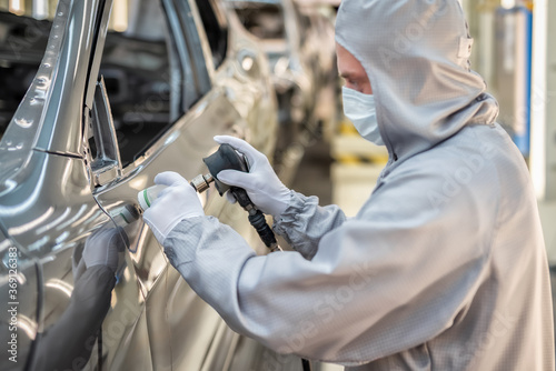 An employee of the car body paint shop with a medical mask on his face grinds the painted surface