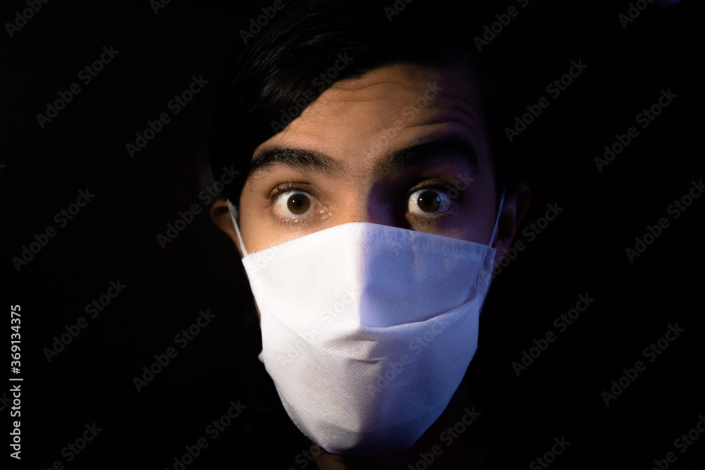 Portrait of a man with a disposable white face mask looking at camera with a surprise face expression. Low key light photography with black background Portrait of a man with a disposable white face mask looking at camera with a surprise face expression. Low key light photography with black background