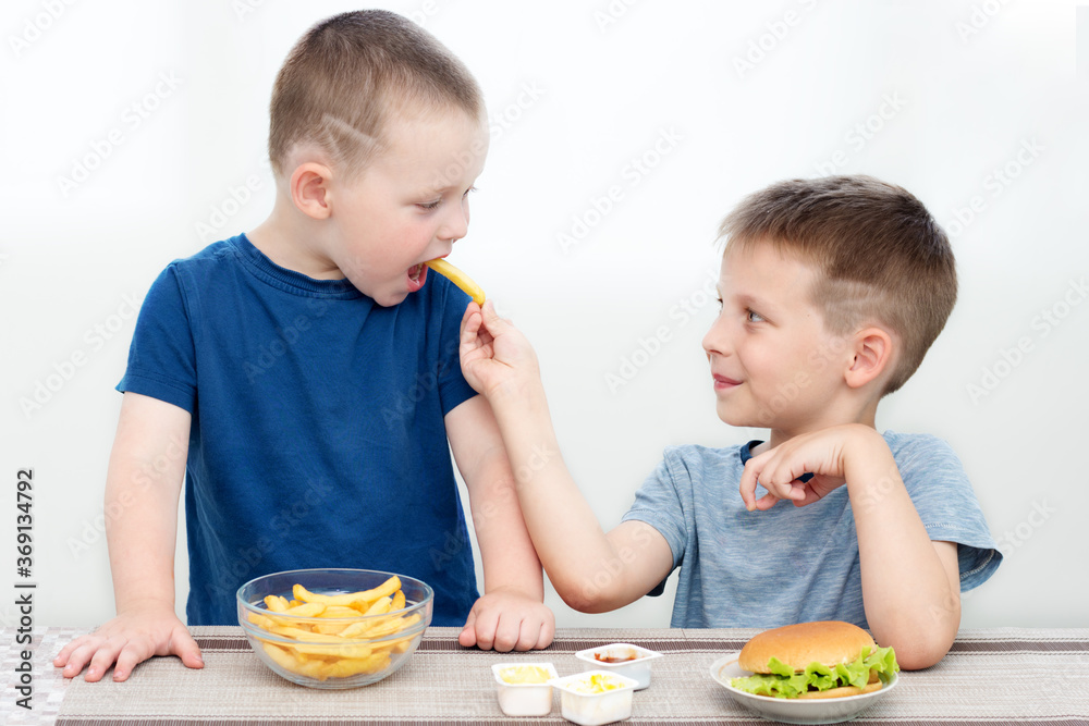 Two happy preschoolers, brothers with an appetite for fast food.One feeds the other with French fries.The food is unhealthy, not suitable for baby food, but delicious,the children are happy,smiling. Two happy preschoolers, brothers with an appetite for fast food.One feeds the other with French fries.The food is unhealthy, not suitable for baby food, but delicious,the children are happy,smiling.