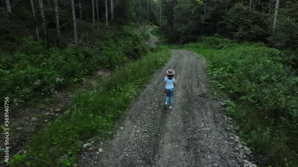 Drone camera follows young woman in hat walks on road near wild forest, enjoys nature. Dreamy walkthrough. Wanderlust explorer and freedom lover in natural surroundings