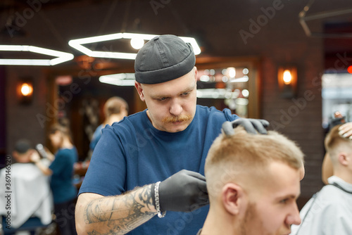 Focused professional barber working with hair clipper, making trendy haircut for young man. Focus on a tattooed man