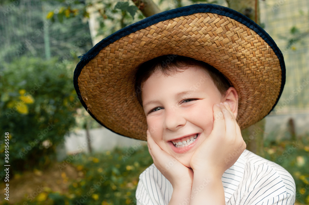 funny pre-school boy in a straw hat and a rustic white shirt sits under an Apple tree in the home garden and smiles, close-up portrait of child
