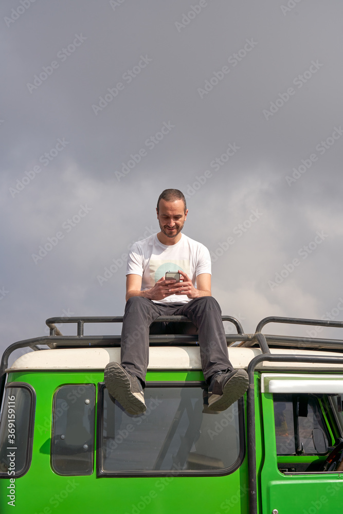 Man visiting Bromo volcano by jeep and sitting on the top of the car using a mobile phone