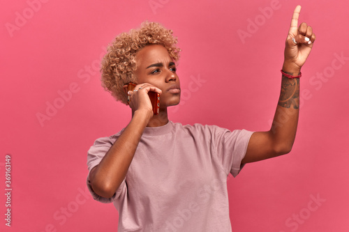 Young dark skinned female listens intently to someone on the smart phone and makes an important decision by raising her index finger up pising over pink background. Advertising for mobile operators. photo