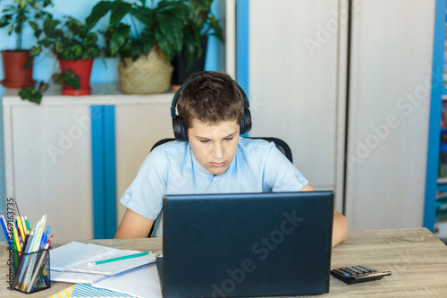 Cute young boy in blue shirt sitting behind desk in his room next to laptop and study. Teenager in earphones makes homework, listening lesson. Home, distance education, self education by kids.