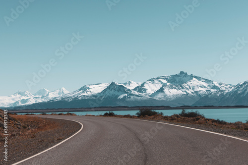 Snowy mountains and large lake after a curve to the left in an asphalt road on a sunny day with blue sky in Patagonia, Chile.
