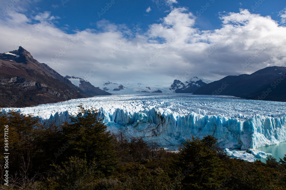 Ice cliff wall behind trees and vegetation in a valley surrounded by snowy mountains at Perito Moreno Glacier in Argentina. Ice cliff wall behind trees and vegetation in a valley surrounded by snowy mountains at Perito Moreno Glacier in Argentina.