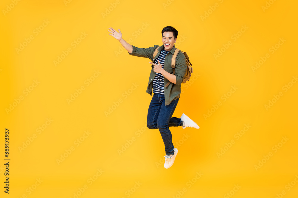 Excited smiling Asian tourist man with backpack jumping and pointing hands to empty space aside on isolated yellow studio background