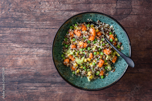 Cold quinoa vegetable salad in a ceramic green bowl, with a black spoon, on a rustic wood background
