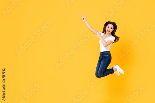 Happy smiling young Asian girl jumping with hands pointing up isolated on yellow studio background with copy space