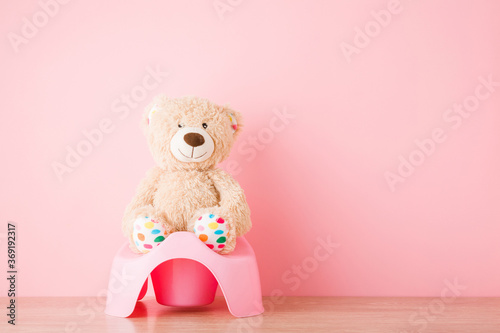 Brown teddy bear sitting on baby potty on floor. Front view. Closeup. Empty place for text on light pink wall background. Pastel color.