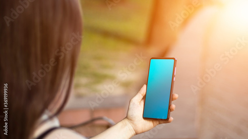 A brunette girl is sitting in the park, holding a mobile phone in her hands. Sunlight, blurred background. lifestyle.