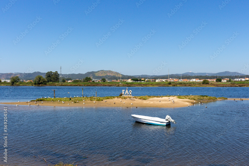 Fão / Esposende / Portugal - July 29, 2020: A small boat moored in the Cavado River estuary on a sunny day. Word FAO made of stone concrete letters. Fão / Esposende / Portugal - July 29, 2020: A small boat moored in the Cavado River estuary on a sunny day. Word FAO made of stone concrete letters.