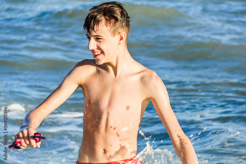 Happy smiling teenager on the background of the sea. A teenager holds swimming goggles in his hand. Splashes from the sea wave. Summer vacation at the sea. Swimming in the sea. Summer tourism.