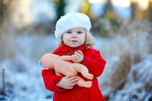 Outdoor winter portrait of little cute toddler girl in red coat and white fashion hat barret. Healthy happy baby child walking in the park on cold day with snow and snowfall. Stylish clothes for kids. photo