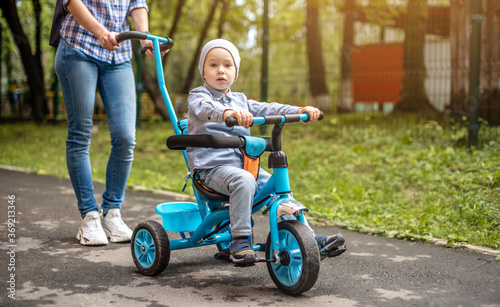 Young mother is pushing a child's tricycle with a toddler boy on a walk. Concept of learning to ride a bike and having fun with your family photo