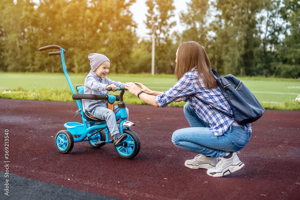 Happy toddler boy is successfully going to his mother on a children's tricycle. Concept of learning to ride a bike and having fun with your family Happy toddler boy is successfully going to his mother on a children's tricycle. Concept of learning to ride a bike and having fun with your family