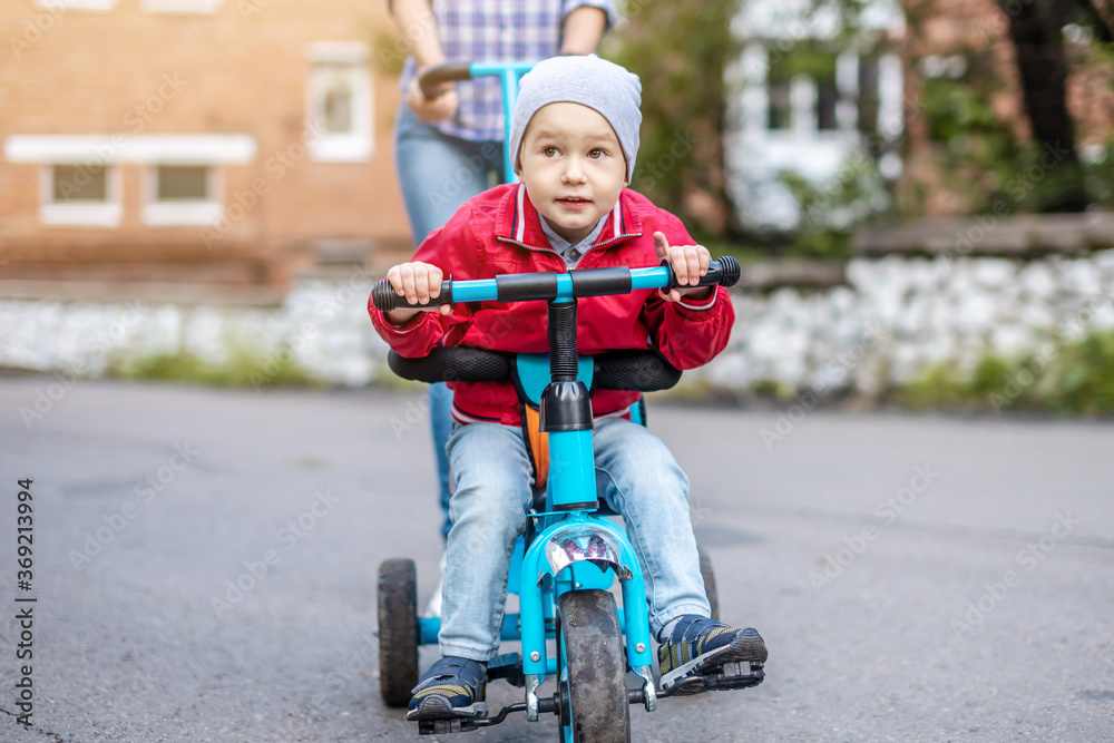 Young mother is pushing a child's tricycle with a toddler boy on a walk. Concept of learning to ride a bike and having fun with your family Young mother is pushing a child's tricycle with a toddler boy on a walk. Concept of learning to ride a bike and having fun with your family