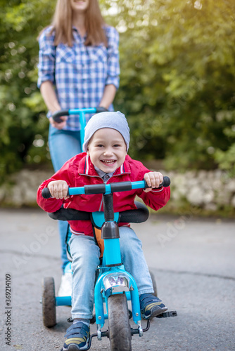 Young mother is pushing a child's tricycle with a toddler boy on a walk. Concept of learning to ride a bike and having fun with your family