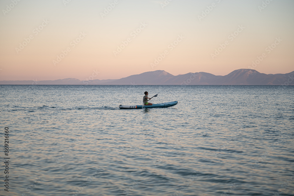 Fototapeta premium Beautiful view of young boy sitting on sup board paddling on calm evening sea
