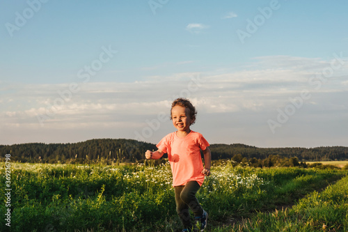 Little curly boy in orange t-shirt running in a meadow, sunset light. Beautiful landscape in summer, very light and happy scene.