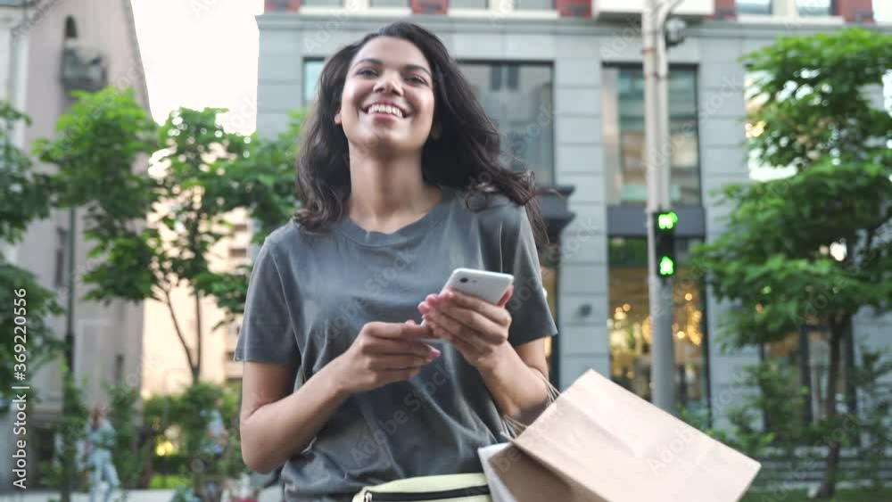 Smiling happy young adult African American ethnic girl shopper customer walking on city street holding shopping bags and smart cell phone using mobile fashion sale ecommerce app near shop mall store.