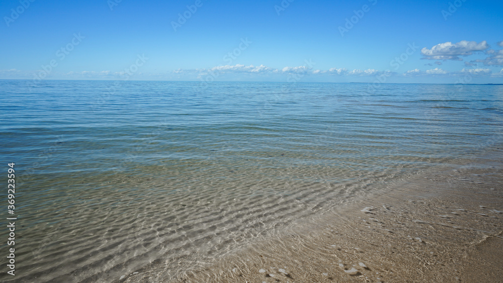Gentle waves wash over sand ripples visible through the clear water. Blue sea stretches to the horizon. Clouds on the horizon. Burrum Heads, Queensland, Australia.