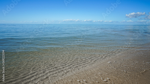 Gentle waves wash over sand ripples visible through the clear water. Blue sea stretches to the horizon. Clouds on the horizon. Burrum Heads  Queensland  Australia.