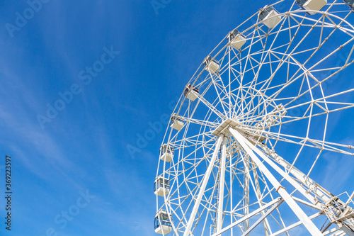 Big ferris wheel with white cabins in an amusement park against a blue sky during the sunny day