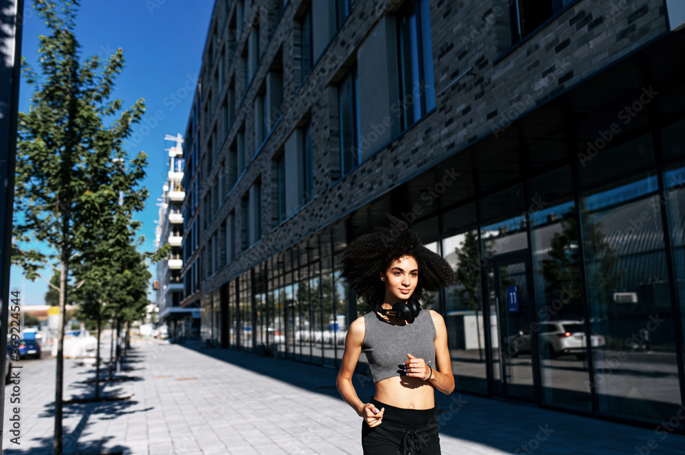 Midshot of attractive African-American woman jogging on the street. A woman with an afro hairstyle with a headphones on the neck is running among city landscape