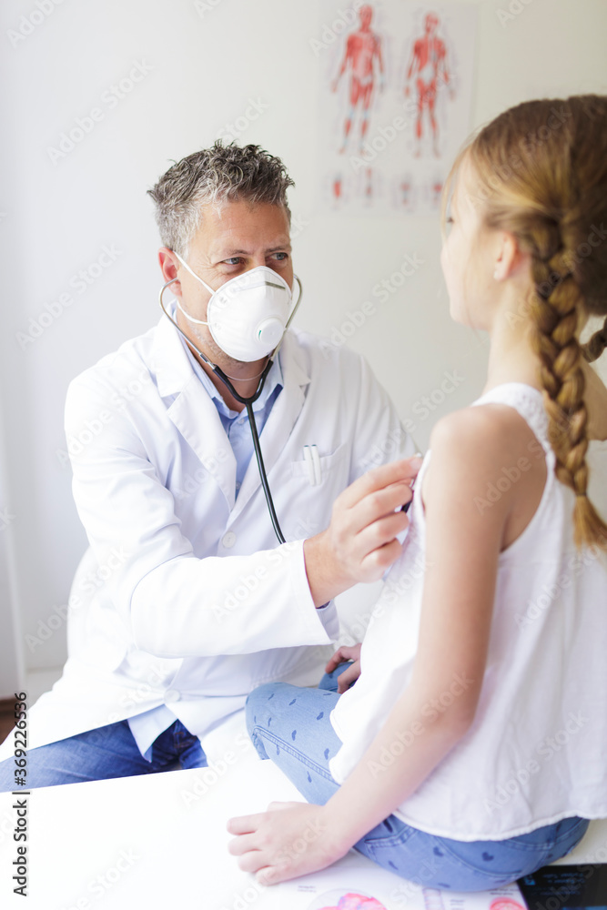 good looking male doctor with three-day beard and nose and mouth mask examines young girl with a stethoscope