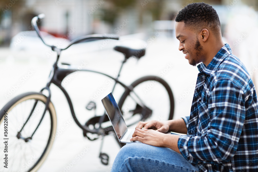 Black man using laptop, sitting in the city with bike Black man using laptop, sitting in the city with bike