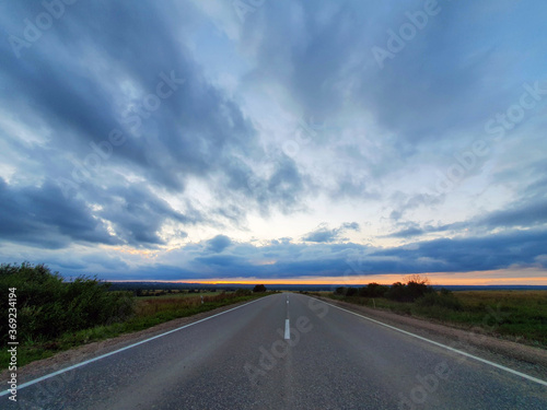an asphalt road (stone grey) surrounded by nature, fields, forests and an unusually colorful sky at sunset. Twilight