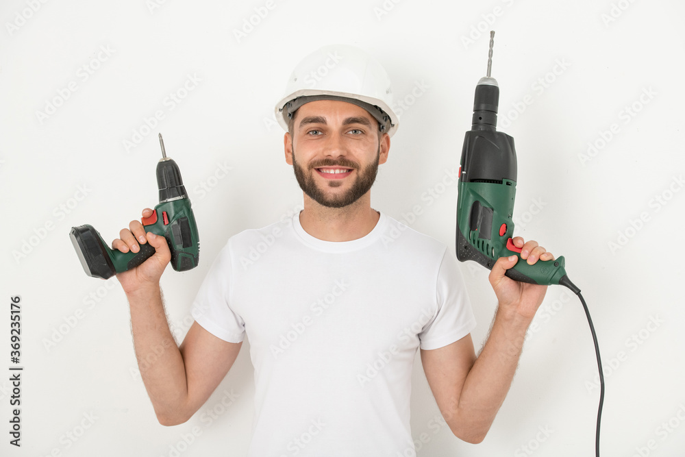 Portrait of smiling young bearded repairman in hardhat holding cordless screwdriver and drill against white wall Portrait of smiling young bearded repairman in hardhat holding cordless screwdriver and drill against white wall