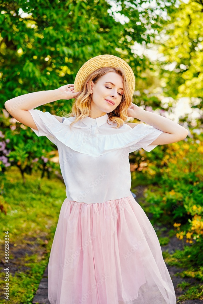 Obraz premium Gorgeous young woman in hat posing in a blooming garden. Beautiful girl dressed in pink skirt and white blouse. Sensual woman with perfect skin and with natural makeup. fashion and style concept.