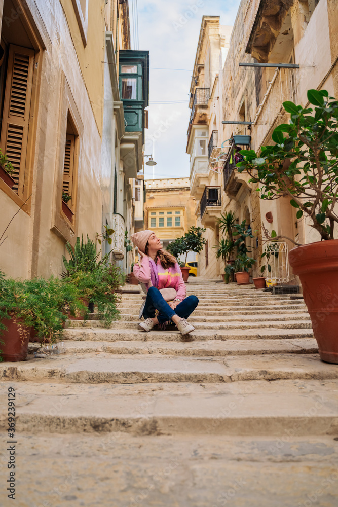 a young woman sits on the steps in a European town with narrow streets, plants in pots along the street, admires the view, looks up. girl in a hat, fall or spring. tourism.